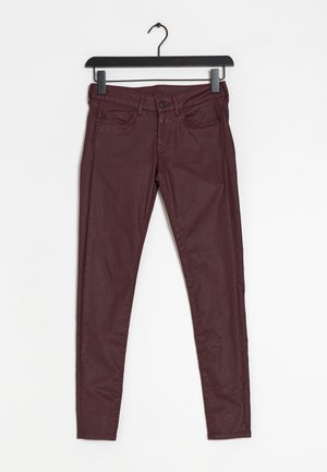 Jean skinny bordeaux avec une texture lisse, poches avant et fermeture éclair, présentée sur un cintre noir sur fond blanc.