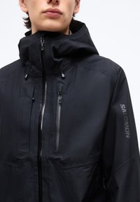 Νεαρό άτομο που φορά μαύρο εξωτερικό μπουφάν Salomon με ψηλό γιακά και πολλαπλές τσέπες με φερμουάρ σε λευκό φόντο.