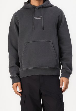 Mann trägt einen dunkelgrauen Hoodie mit Fronttasche und dem Text "SAMSØE SAMSØE COPENHAGEN", kombiniert mit schwarzen Cargohosen.