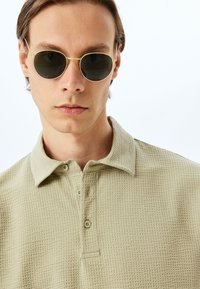 Polo shirt beige chiaro con un motivo testurizzato, dotato di colletto e zip a tre bottoni. Il indossatore indossa occhiali da sole scuri e rotondi.