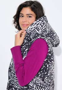Zwart-witte gecamoufleerde puffer vest met een grote capuchon, gedragen over een effen paarse longsleeve shirt, waardoor textuur en contrast benadrukt worden.