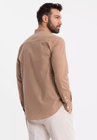 Chemise marron à manches longues avec un col pointu, des poignets boutonnés et un ourlet arrondi. Texture lisse et design simple, présentée de dos.