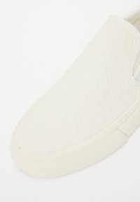 Hvit slip-on sko med et teksturert snøremønster, gummisåle og elastiske detaljer ved åpningene for enkel påkledning.