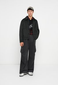 Grijze button-up jas over een zwart graphic t-shirt, gecombineerd met losse, donkere cargobroek en witte sneakers met zwarte strepen; lichte pet.