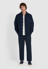 Mężczyzna z kręconymi włosami, ubrany w granatową koszulę zapinaną na guziki na białej koszulce, granatowe spodnie i granatowe tenisówki, stojący na tle jednolitego tła.