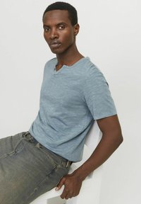 Lyseblå kortærmet t-shirt med en tekstureret overflade og V-udskæring, parret med mørkegrå jeans. Modellen sidder på en stol.