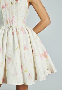 Vestido de estampado floral, confeccionado en una tela ligera. Cuenta con un corpiño ajustado, falda plisada y un fondo crema con acentos en rosa y verde.