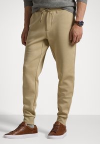 Mężczyzna ubrany w beżowe spodnie dresowe, szare skarpetki, brązowe skórzane sneakersy, szary sweter oraz zegarek z niebieską tarczą na lewym nadgarstku na jednolitym tle.