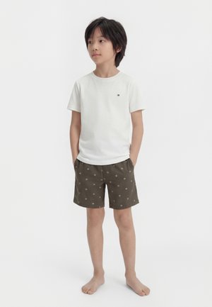 Chlapec stojaci na boso, oblečený v bielom tričku s krátkym rukávom a tmavých vzorovaných krátkych nohaviciach, s rukami vo vreckách na bielom pozadí.