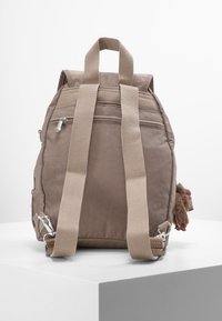 Beige stof rygsæk med justerbare brune stropper, to lynlåsede lommer og et blødt element fastgjort til siden. Kompakt og rektangulær form.