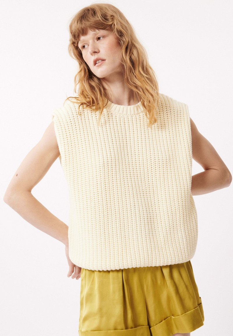 Pull sans manches de couleur crème en tricot avec un motif texturé, encolure ronde et coupe décontractée, associé à un short plissé vert olive.