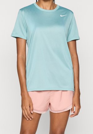 Femme portant un t-shirt de sport à manches courtes Nike bleu clair et un short de course rose clair, debout devant un fond uni.