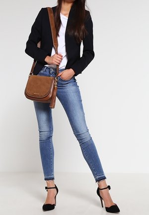 Mujer con blazer negro, camiseta blanca, jeans ajustados azules, tacones altos negros y un bolso cruzado de cuero marrón con detalle de flecos.