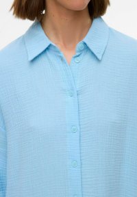 Light blue button-up shirt made of textured fabric with a collared neck and six matching buttons down the front.