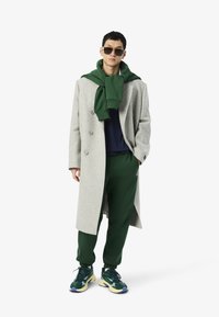 Manteau en laine grise avec des boutons croisés, sweat à capuche vert drapé au cou, chemise bleu marine, joggeurs verts et baskets de sport multicolores.