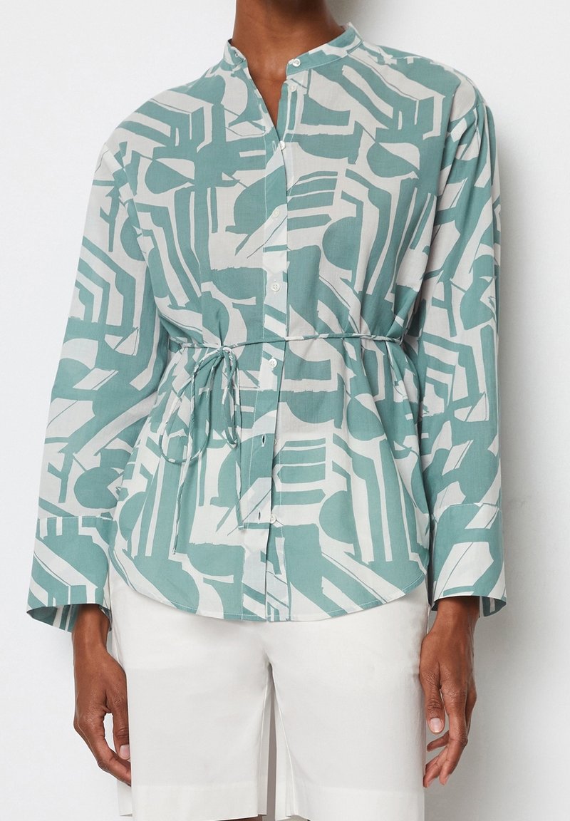 Chemise à manches longues boutonnée avec un motif géométrique abstrait en bleu sarcelle et blanc, nouée à la taille, associée à un short blanc.