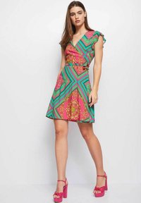 Vestido de manga corta en un patrón rosa, verde y dorado, con escote en V, cintura ceñida con cinturón y falda acampanada. Combinado con sandalias de tacón bloque en color rosa.