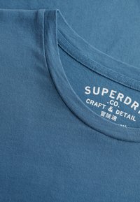 T-shirt bleu à col rond avec un texte de marque blanc imprimé partiellement visible « SUPERDRY » et « CRAFT & DETAIL » à l'intérieur du col.