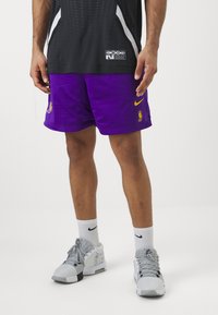 Vijolične košarkarske kratke hlače iz mrežastega materiala z rumenimi detajli, opremljene z NBA logotipom in Nike blagovno znamko. Oblečene so z sivimi supergami in belimi Nike nogavicami.