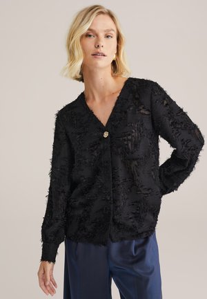 Cardigan nero in tessuto texture con scollatura a V, dotato di un bottone centrale dorato. Maniche lunghe con dettagli frangiati, indossato sopra pantaloni in satin blu navy.