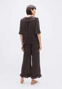 Femme debout pieds nus, portant un ensemble de pyjama noir à pois avec un ourlet froncé et un large col, vue de dos sur un fond uni.