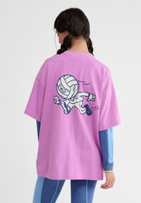 Lyserød oversized bomulds t-shirt med en blå grafisk illustration af en karakter, der løber. T-shirten har korte ærmer og en rund halsudskæring.