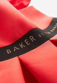 Tissu rouge avec un élastique noir affichant un texte brodé en or « BAKER BY TED BAKER » le long du bord.
