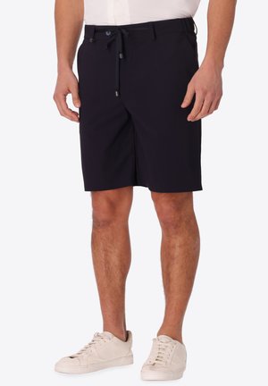 Uomo che indossa pantaloncini neri con coulisse e sneakers bianche, in piedi davanti a uno sfondo chiaro e uniforme.