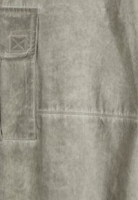 Tessuto grigio chiaro con una texture morbida; presenta una tasca a toppa quadrata e dettagli di cucitura visibili. Design pulito e semplice.