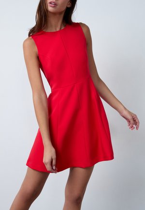 Femme portant une robe rouge sans manches, longueur genoux, avec une taille cintrée, sur un fond clair uni.