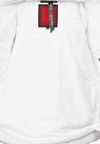 Vit fleece hoodie med en strukturerad yta; har en röd etikett på framsidan och ett svart band med varumärkeslogotyp i närheten av halsringningen.