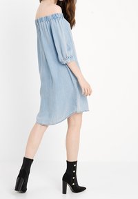 Světle modré šaty z denimu s odhalenými rameny, volnými rukávy, elastickým výstřihem a zakřiveným lemem, v kombinaci s černými kotníkovými botami s zlatými knoflíčky.