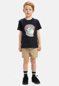 Camiseta de algodón negra con un gráfico colorido de surf, combinada con pantalones cortos beige. Lleva zapatillas negras y calcetines negros con rayas blancas.