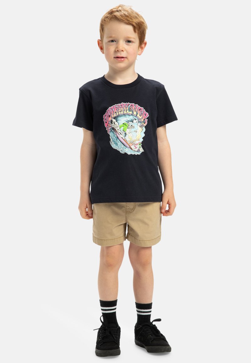 Camiseta de algodón negra con un gráfico colorido de surf, combinada con pantalones cortos beige. Lleva zapatillas negras y calcetines negros con rayas blancas.