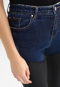 Vaqueros de denim con un lavado oscuro y una textura suave, cintura de altura media, diseño clásico de cinco bolsillos y detalles de cuero negro en la parte inferior de la pierna.