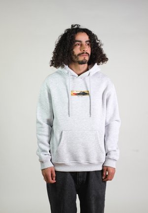 Man met krullend haar die een lichtgrijze hoodie draagt met een kleine afbeelding van een zwarte auto met vlam op de borst, staand tegen een effen achtergrond.