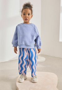 Hellblaues Sweatshirt mit Raglanärmeln, kombiniert mit rosa und blauen Hosen mit Wellenmuster. Das Model trägt weiße Sneakers.