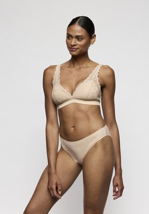 Kobieta w beżowym koronkowym biustonoszu i dopasowanych majtkach z gładkiego materiału, stojąca na tle jednolitego, jasnoszarego tła.