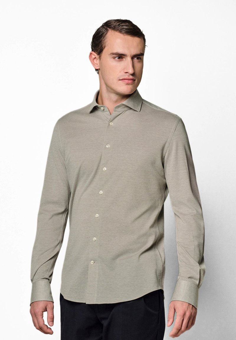 Homme portant une chemise beige à manches longues boutonnée et un pantalon noir, regardant vers la droite sur un fond blanc uni.