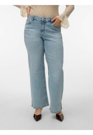 Vrouw die lichtblauwe jeans met een hoge taille en wijde pijpen draagt, een kanten top met lange mouwen en puntige bruine schoenen, met haar handen in de voorzakken.