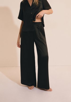 Femme portant un pantalon ample en satin noir et une chemise courte à manches courtes assortie, soulevant légèrement la chemise pour dévoiler la taille.