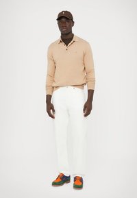 Beige langermet polo-skjorte, hvite jeans og flerfargede joggesko. Tilbehør inkluderer en brun caps. Enkel, moderne antrekk.