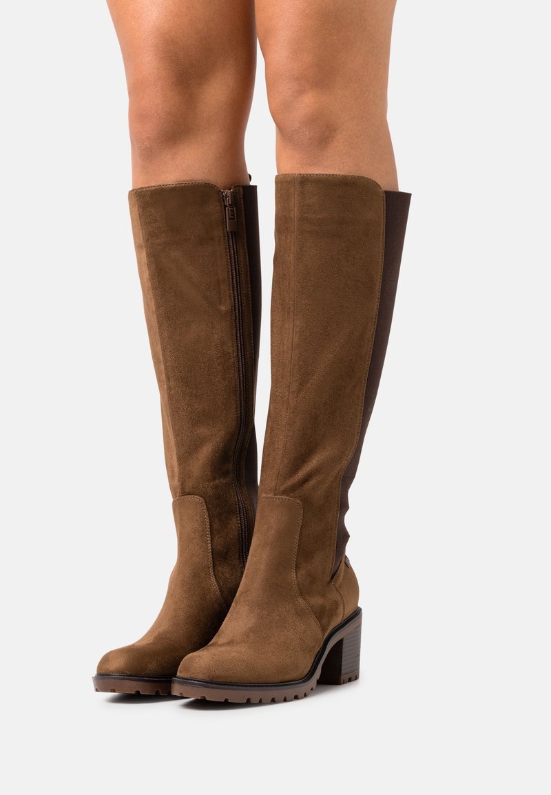 Paire de bottes montantes en suède marron avec talons blocs et fermetures éclair latérales portées sur des jambes nues, sur un fond uni.