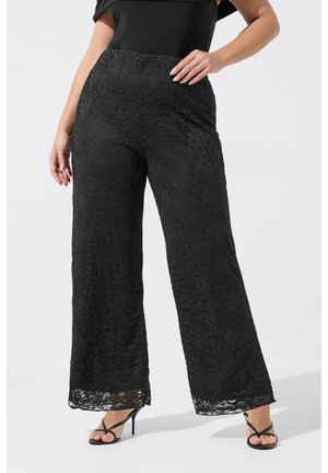 Kvinde iført sorte, sortblonde bukser med høj talje og vide ben, kombineret med sorte, remformede højhælede sandaler, poserer med den ene hånd på hoften.