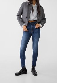 Mujer vestida con un blazer corto gris, una sudadera con cremallera gris claro, unos vaqueros ajustados de cintura alta en azul, calcetines negros y unos mocasines negros.