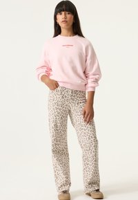 Mujer con sudadera rosa claro con el texto "FIELD OF DREAMS" y pantalones beige con estampado de leopardo, de pie con una mano en el bolsillo.