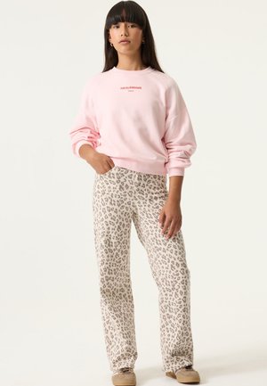 Femme portant un sweat-shirt rose clair avec le texte "FIELD OF DREAMS" et un pantalon beige à imprimé léopard, debout avec une main dans la poche.