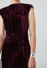 Robe en velours cramoisi à rayures, avec un détail de taille noué. Un bracelet de poignet en argent métallique met en valeur le design.