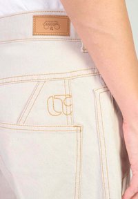 Pantalons en denim beige avec des coutures contrastées, une étiquette en cuir sur la taille et un détail de poche brodé unique.