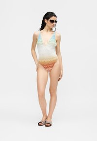 Γυναίκα που φοράει μονόχρωμο ολόσωμο μαγιό με βαθύ V λαιμόκοψη σε πολύχρωμο διαβαθμισμένο σχέδιο, μαύρα γυαλιά ηλίου και σανδάλια τύπου slide με μοτίβα, στέκεται μπροστά σε απλό φόντο.